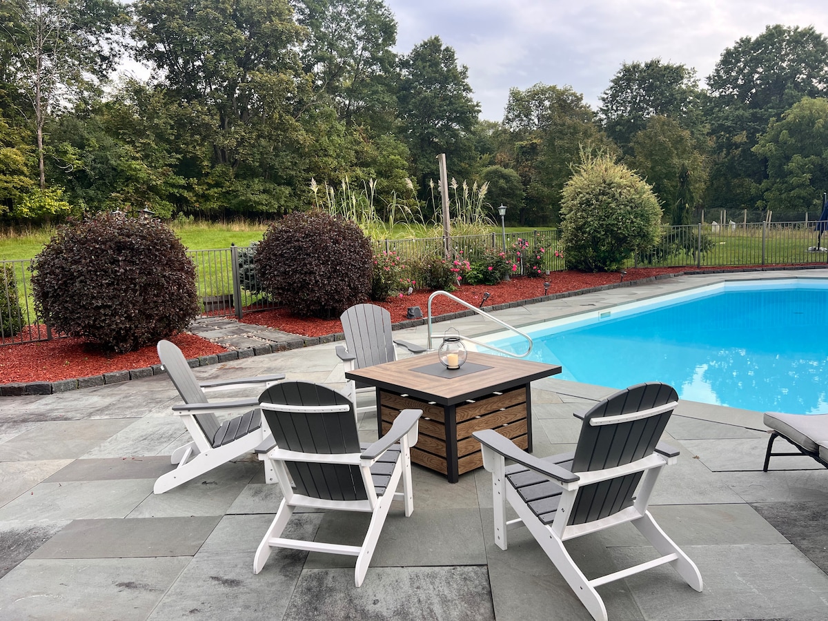 The pool terrace at Thee Wild Lux — Adirondack chairs, fire pit table, lush summer gardens